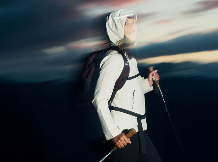 A person in hiking gear, including a white jacket and hood, smiles while holding trekking poles. The blurred twilight sky suggests movement and adventure.