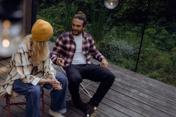 Two young adults in casual outdoor clothing sit on chairs in front of a wooden hut in the forest