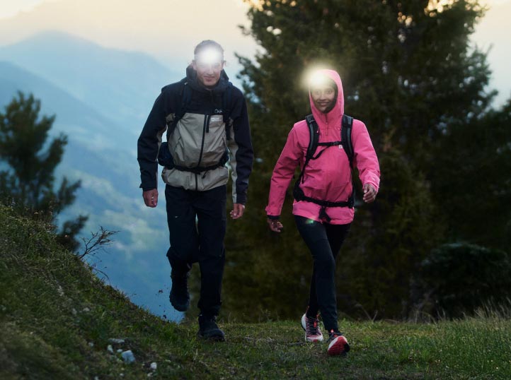 Two hikers with headlamps walk on a forest trail at dusk. They wear jackets, one black and gray, the other pink.